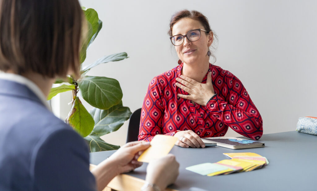 Coach Susanne an einem Tisch sitzend im Gespräch mit einer Klientin, vor sich farbige Karten und ein Notizheft während einem Coaching für Schulleitungen. Susanne legt die Hand auf die Brust und lächelt die Klientin an