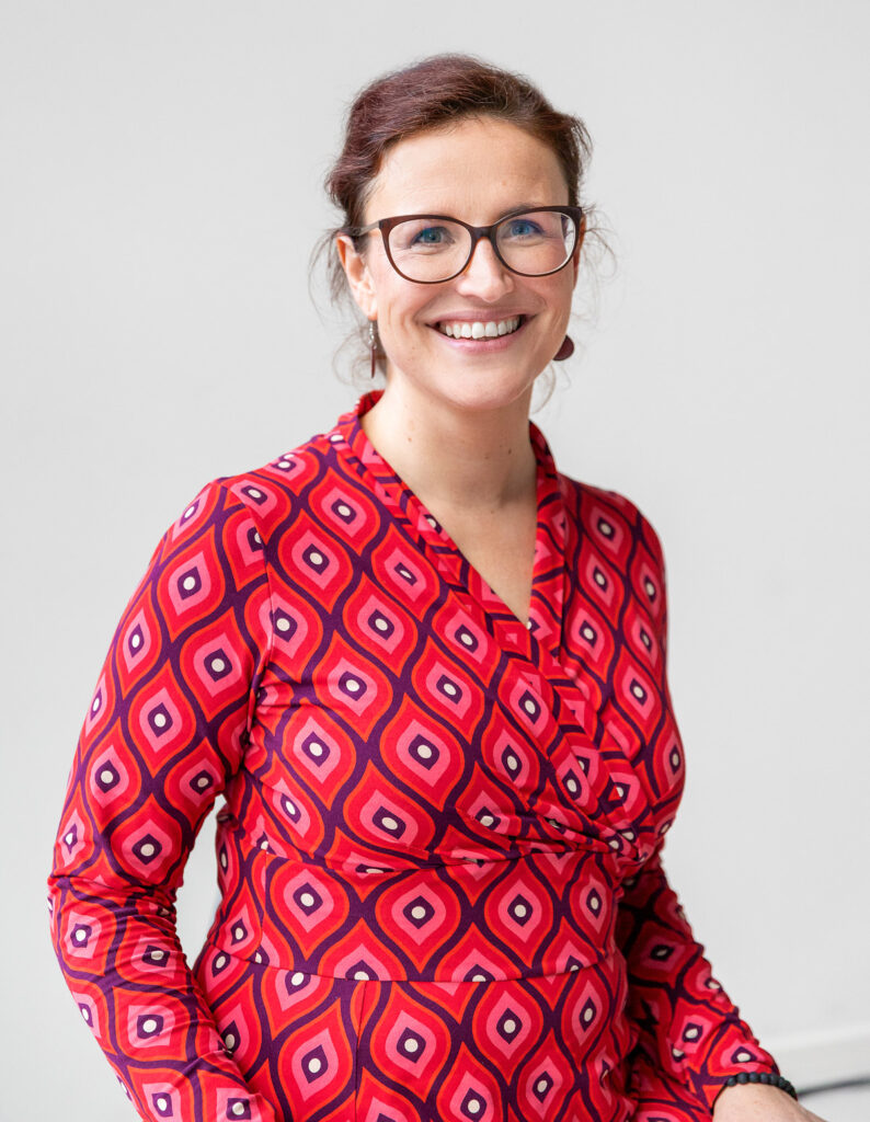 Portrait von Susanne, in die Kamera lächelnd, vor einer hellen Wand, mit einem rot-gemusterten Kleid